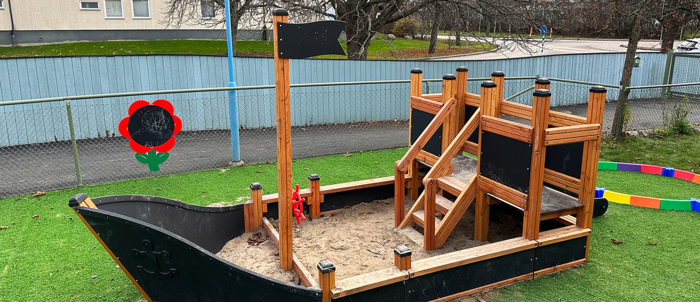 legeplads med stort, nedgravet skib fyldt med sand placeret på kunststofgræs